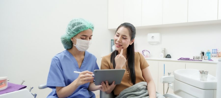 Asian adult female patient in dental chair pointing to cheek during dental consultation with dental professionals collaborating in office